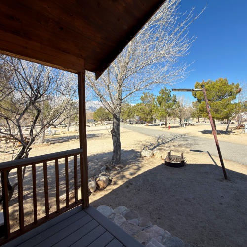 A desert porch scene with a wooden railing, steps, bare trees, a dirt yard, and a bright blue sky in a quiet neighborhood.