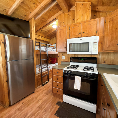 A cozy wooden kitchen with stainless fridge, microwave above a gas stove, oak cabinets, a sink, and an open shelf pantry beside the fridge.
