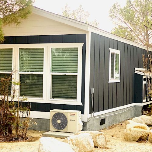 A small, modern house with dark exterior siding, large windows, an air conditioning unit outside, surrounded by trees and rocks.