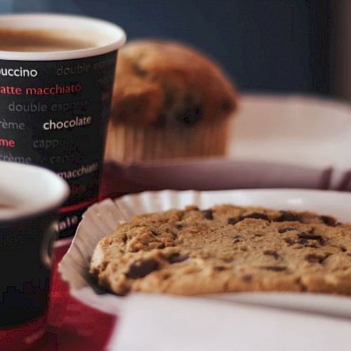 A coffee with text on the cup, a cookie on a plate, and an out-of-focus muffin in the background are on a table.