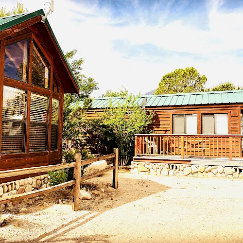 A scenic image of two wooden cabins with green roofs, large windows, a porch, and a dirt pathway, set against a backdrop of trees and a blue sky.