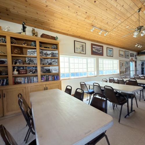 A bright, rustic classroom with long tables, metal chairs, wooden cabinets full of shelves and memorabilia along the left, and framed photos on the walls under a wooden ceiling.