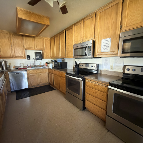 A kitchen with wooden cabinets, stainless steel appliances, a fridge, dishwasher, microwave, and stove; a ceiling fan and papers on the wall.