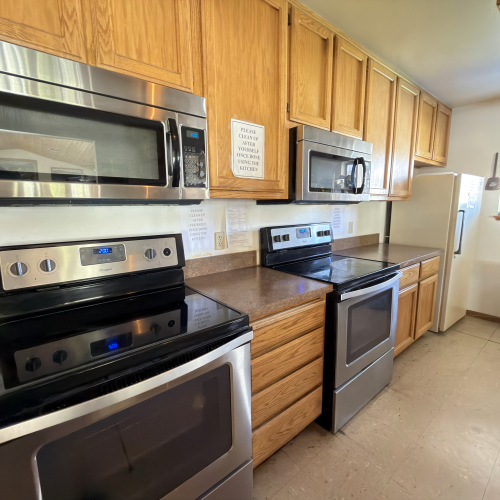 A kitchen with wooden cabinets, stainless steel appliances (microwaves, oven, stove), a white refrigerator, and a sink area, tidy and bright.