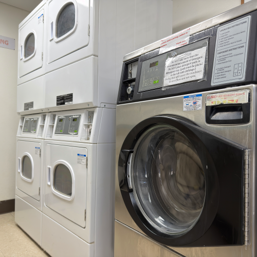 A row of white front-load washing machines stacked in a laundromat next to a large black industrial dryer, with a &ldquo;No Smoking&rdquo; sign visible.