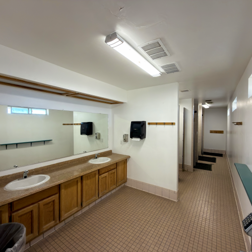 A public restroom with a long wooden vanity, two sinks, mirrors, soap dispensers, and a hallway with stalls in a beige-tiled floor layout.