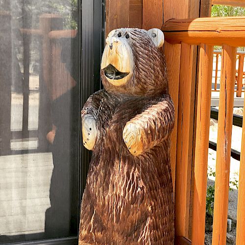 A wooden bear sculpture is standing next to a sliding door and wooden railing on a porch, with its mouth open and arms slightly raised.