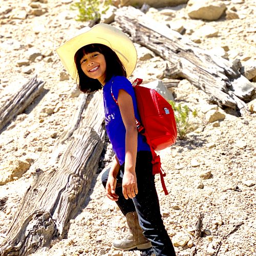 A child with a red backpack and cowboy hat hikes up a rocky slope, smiling brightly under the sun while surrounded by rocks and wood pieces.