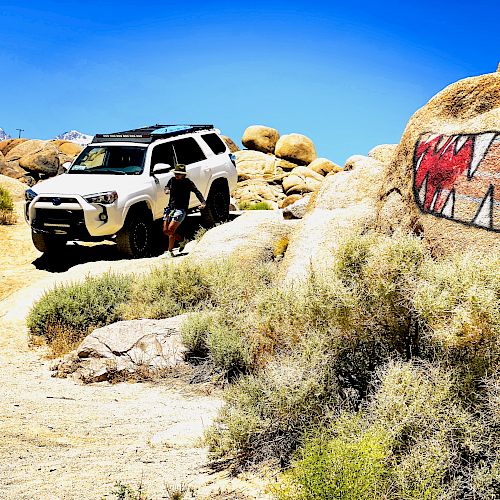 The image shows a white SUV off-roading in a rocky, desert landscape. A rock painted with a mouth and sharp teeth is also visible.