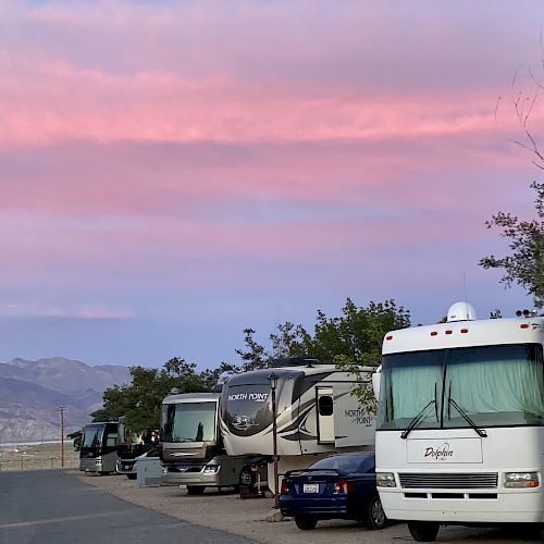 The image shows a row of parked RVs on the side of a road with a stunning pink and purple sky in the background, and mountains in the distance.
