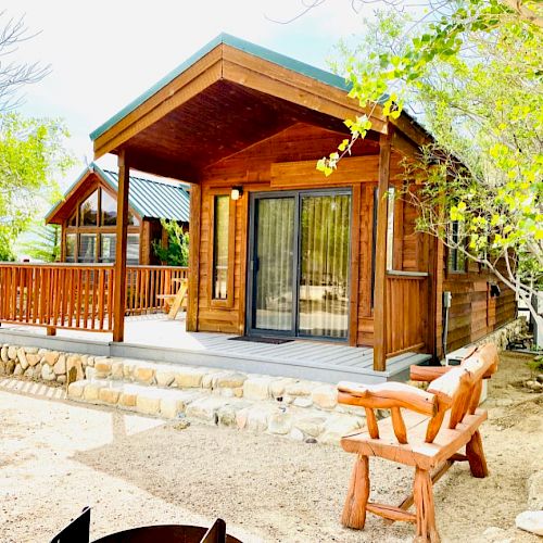 A cozy wooden cabin with a porch and rocking bench surrounded by trees, and another similar cabin visible in the background under a clear sky.