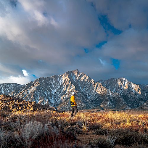 A person in a yellow jacket stands in an expansive landscape with towering, snow-capped mountains under a dramatic cloudy sky.