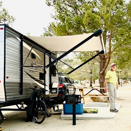 A person stands beside a camper trailer with an awning, parked in a wooded area. The setup includes outdoor gear like a bike and table with supplies.