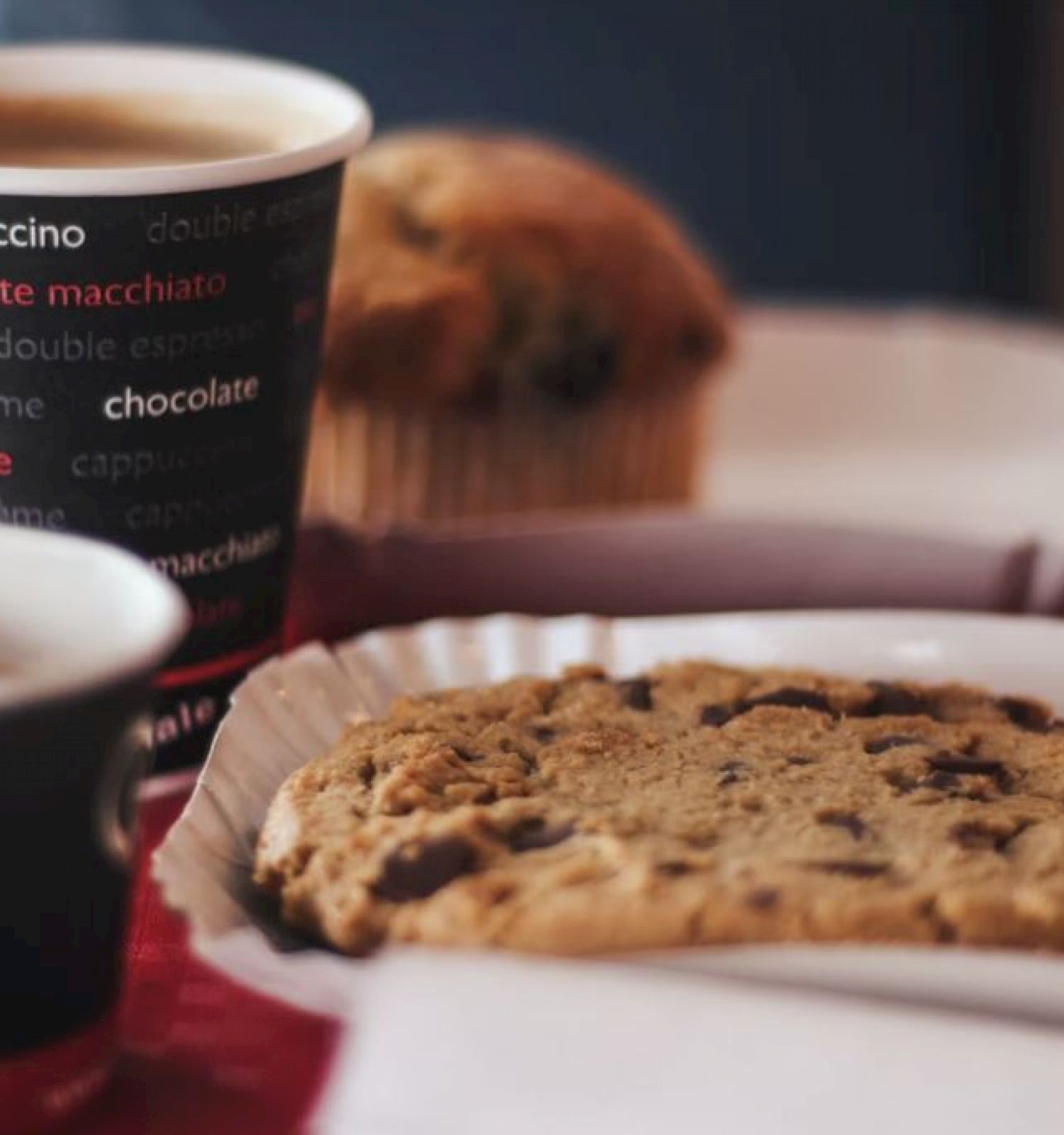 The image shows a paper coffee cup, an espresso cup, a chocolate chip cookie on a plate, and a muffin in the background, placed on a table.