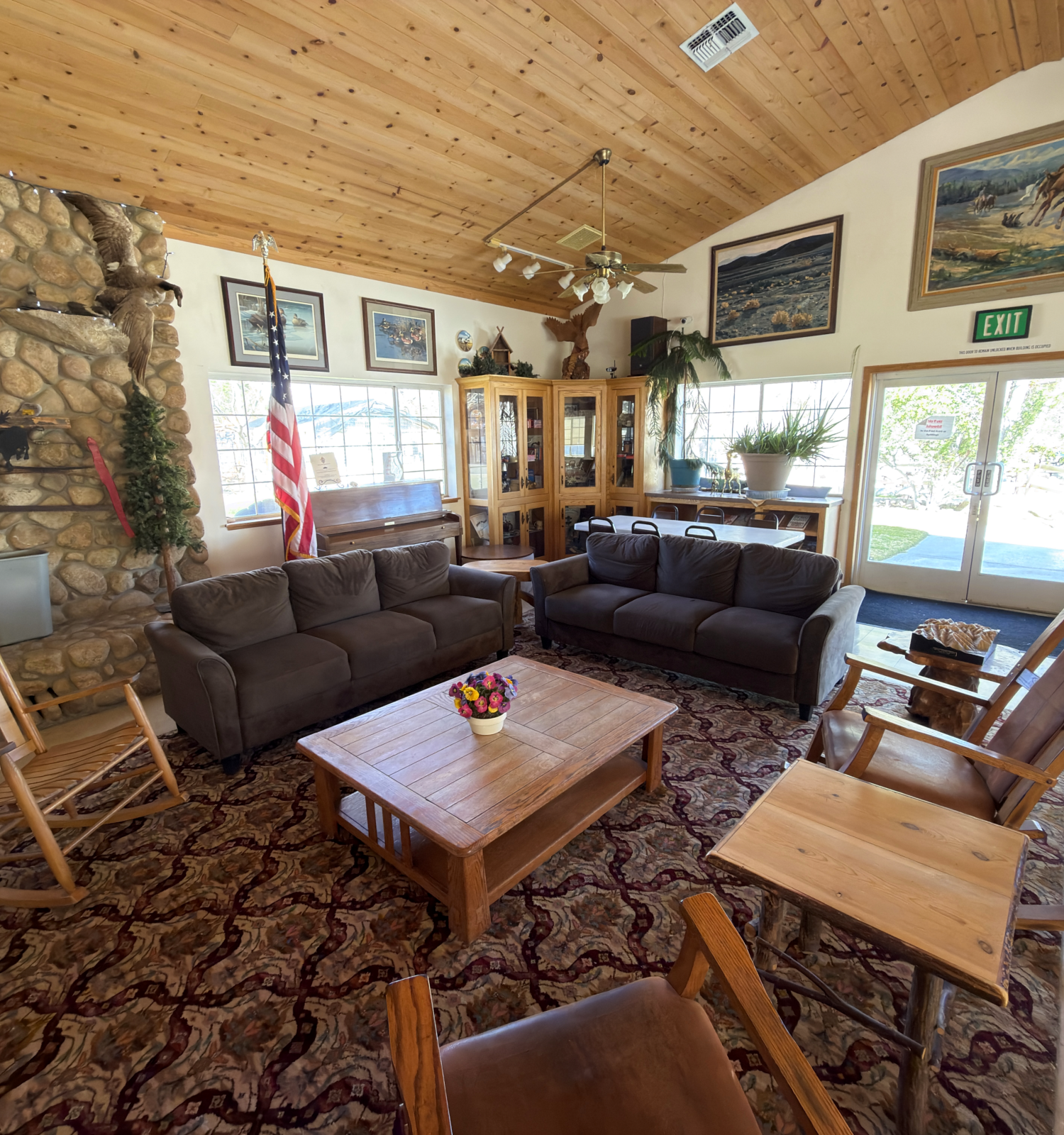 A cozy cabin living room with a stone fireplace, wooden ceiling, brown leather sofas, wooden rockers, a coffee table, and large windows letting in light.