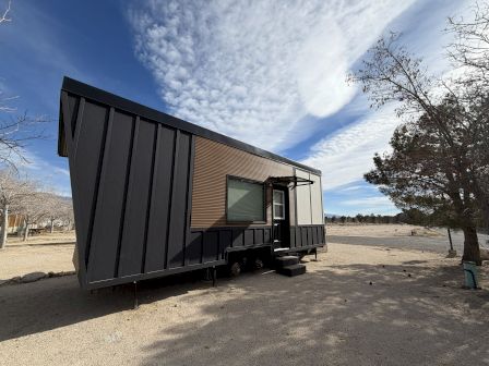 A small, dark metal cabin on wheels sits on a sandy lot with a leafless tree nearby beneath a bright, partly cloudy sky. Top it at 140 characters, always ending the sentence.