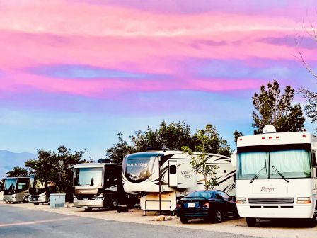 Several RVs parked side by side under a vibrant pink and purple sunset sky, with trees in the background.