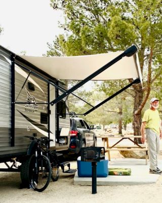 A person standing next to a parked RV with an extended awning in a campsite surrounded by trees, near a table with camping gear.