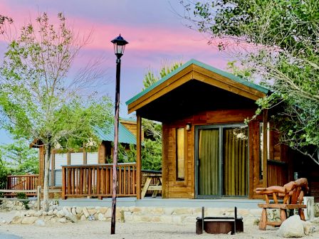 A cozy wooden cabin with a covered porch, surrounded by trees, a bench, and a bright pink sunset sky.