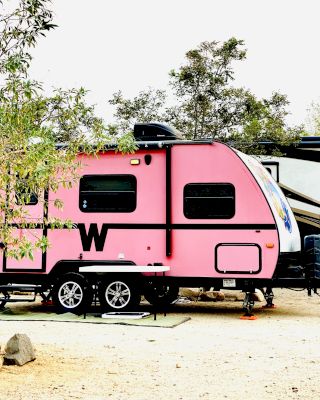 The image shows a pink travel trailer with a large "W" on its side, parked in an outdoor area with trees surrounding it.